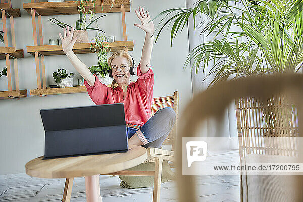 Fröhliche Frau mit erhobenen Händen während eines Videoanrufs zu Hause