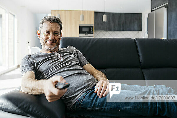 Smiling man holding TV remote while sitting on sofa at living room