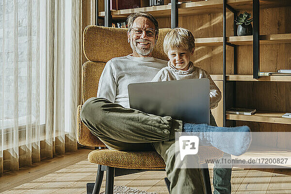 Lächelnder Junge  der einen Laptop benutzt und mit seinem Vater auf einem Stuhl sitzt