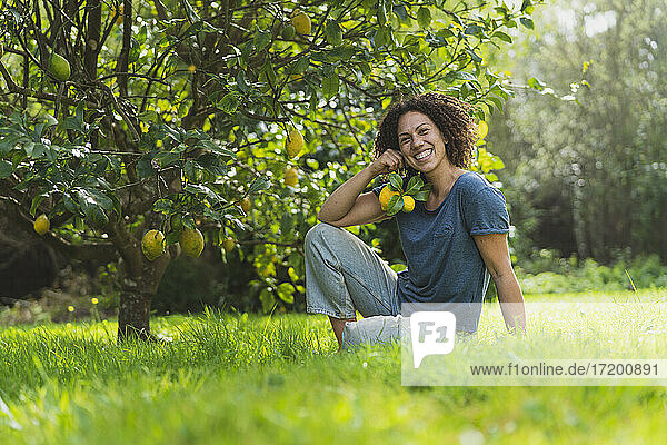 Glückliche Frau hält Zitronen in der Hand und sitzt im Gras unter einem Zitronenbaum