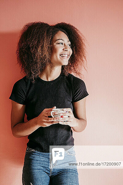 Glückliche Afro-Frau  die eine Kaffeetasse hält und vor einem pfirsichfarbenen Hintergrund wegschaut