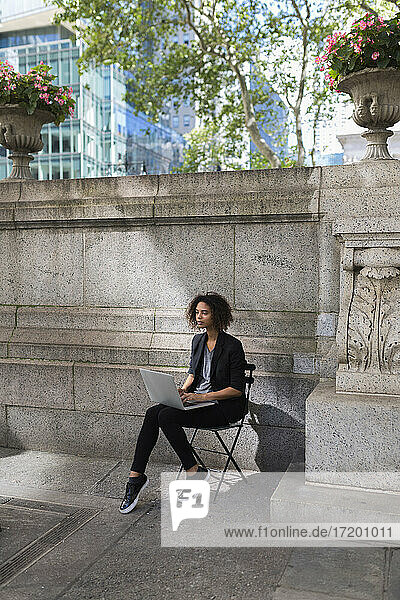 Nachdenkliche Geschäftsfrau  die einen Laptop benutzt und auf einem Stuhl vor einer Wand sitzt