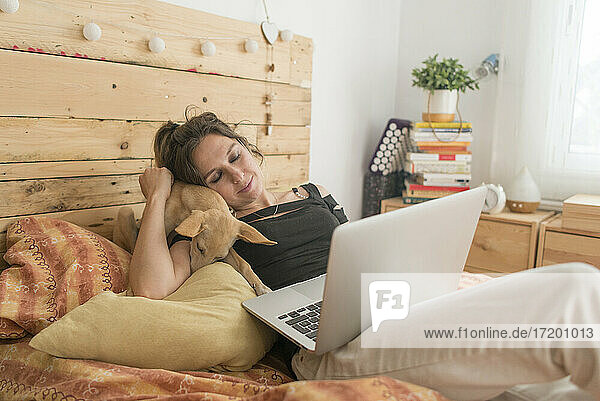 Frau mit Laptop  die ihren Hund umarmt  während sie zu Hause auf dem Bett sitzt