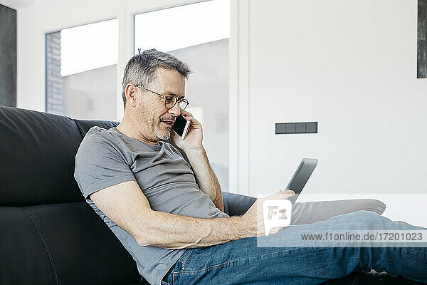 Mature man talking on smart phone while using digital tablet on sofa at living room