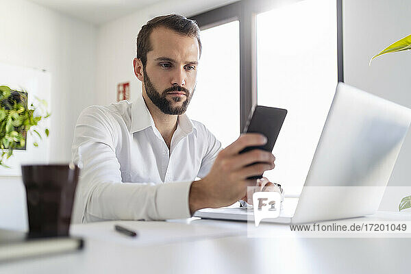 Junger Unternehmer mit Laptop  der ein Mobiltelefon benutzt  während er im Büro sitzt