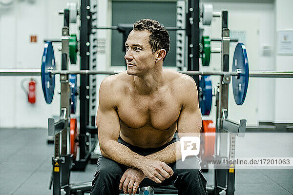 Shirtless male athlete looking away while sitting at bench press in gym