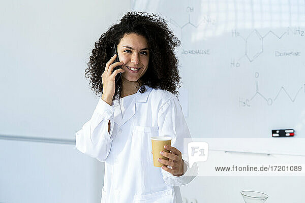 Lächelnde Ärztin mit Kaffeetasse  die in einer Klinik mit einem Mobiltelefon spricht