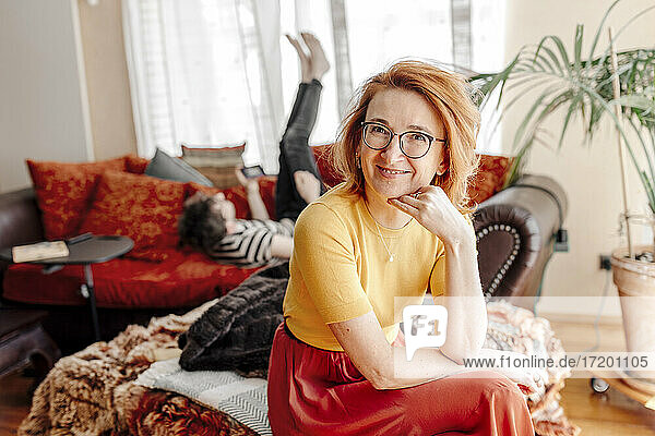 Lächelnde Frau sitzt zu Hause auf der Couch  während sich ihr Sohn auf dem Sofa im Hintergrund entspannt