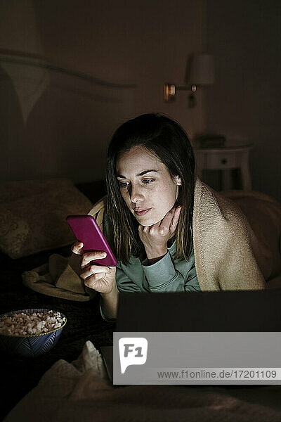 Frau mit Laptop und Popcorn  die ihr Handy benutzt  während sie zu Hause auf dem Bett liegt