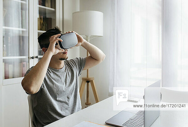 Männlicher Unternehmer  der ein Virtual-Reality-Headset hält  während er mit einem Laptop auf dem Schreibtisch im Heimbüro sitzt