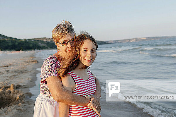 Senior woman embracing granddaughter while standing at beach