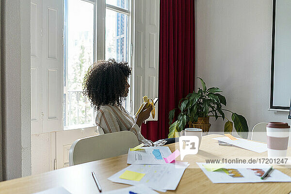 Junge afroamerikanische Unternehmerin  die in der Kaffeepause im Büro ihr Smartphone benutzt und eine Banane isst