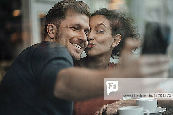 Lächelnder Freund nimmt Selfie mit Freundin durch Handy beim Sitzen im Café