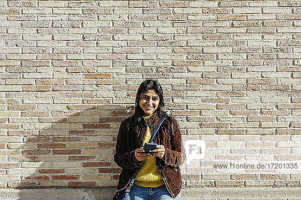Lächelnde Frau mit Umhängetasche  die ihr Mobiltelefon gegen eine Backsteinmauer hält
