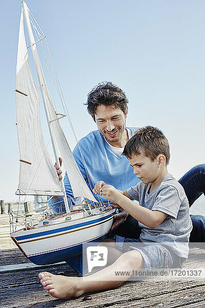 Boy repairing toy sailboat while sitting by father on pier