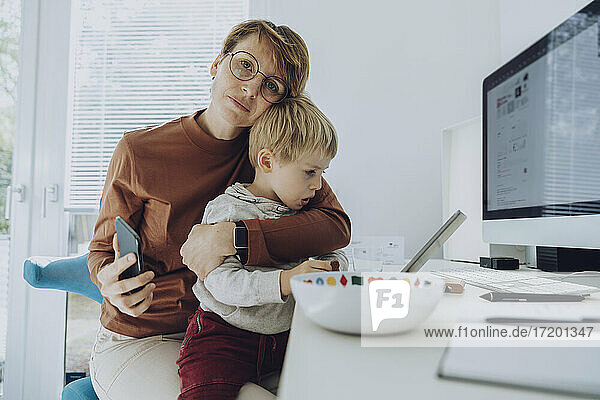 Liebevolle Mutter  die ihren Sohn umarmt  während sie im Büro zu Hause auf ein digitales Tablet schaut