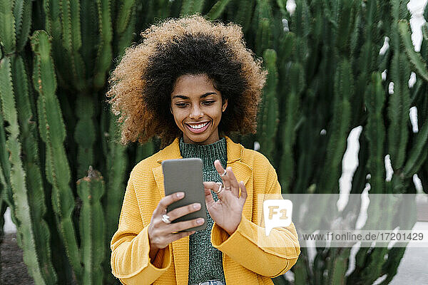 Smiling Afro woman using smart phone while standing against cactus plants