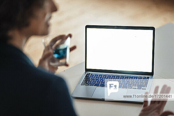 Woman holding glass while gesturing sitting in front of laptop screen