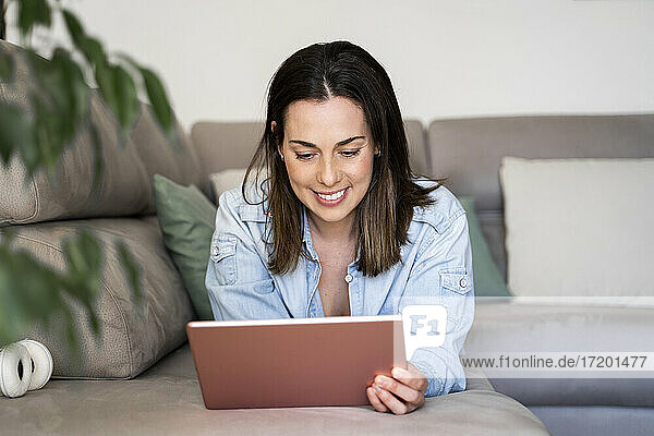 Lächelnde Frau  die ein Tablet benutzt  während sie sich auf dem Sofa zu Hause entspannt