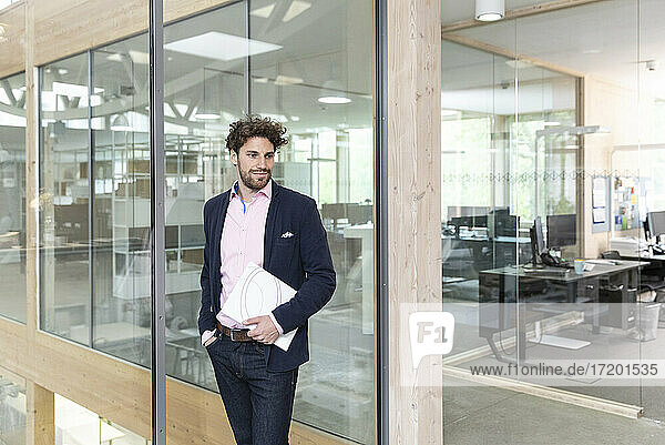 Thoughtful young male entrepreneur holding document while leaning on glass wall at office