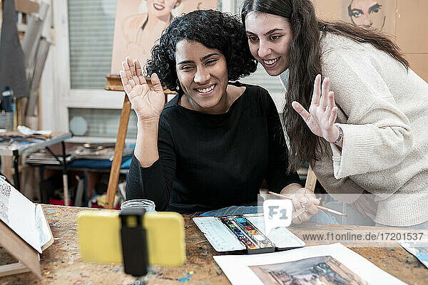 Fröhliche junge Malerinnen  die während eines Videogesprächs im Kunstatelier mit ihrem Smartphone winken