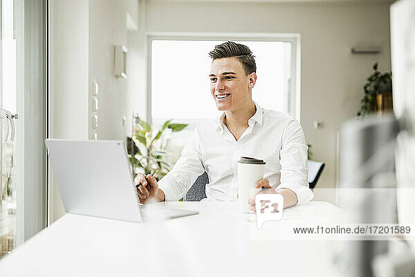 Lächelnder Geschäftsmann mit Einweg-Kaffeebecher und Laptop am Schreibtisch sitzend