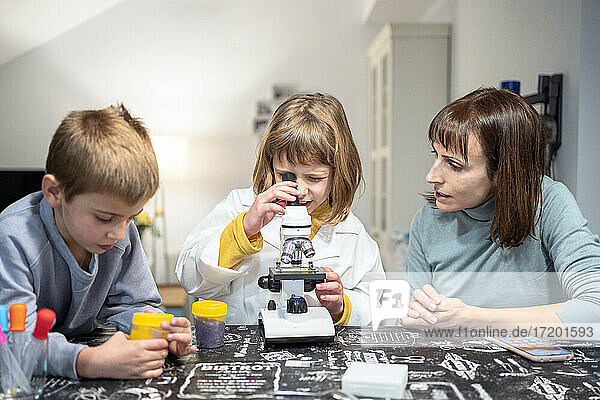 Mädchen schaut durch ein Mikroskop inmitten von Bruder und Mutter zu Hause