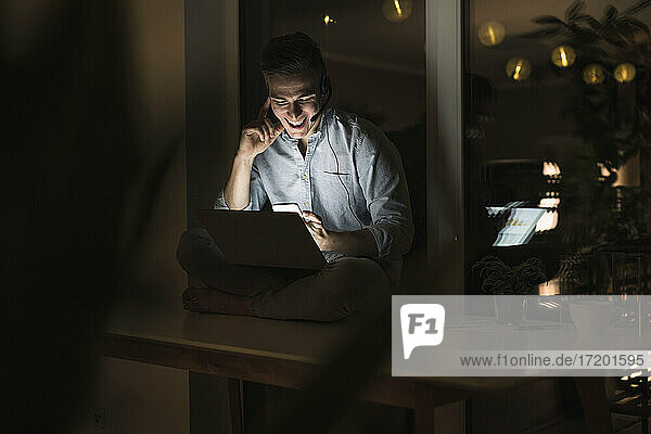 Smiling businessman with laptop using smart phone while sitting on table in living room