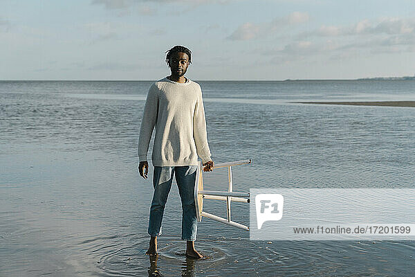 Junger Mann mit weißem Hocker steht am Strand gegen den Himmel