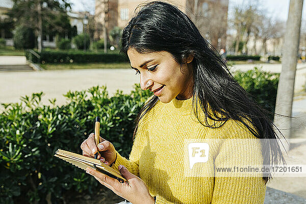 Lächelnde Frau schreibt in einem öffentlichen Park in ihr Tagebuch
