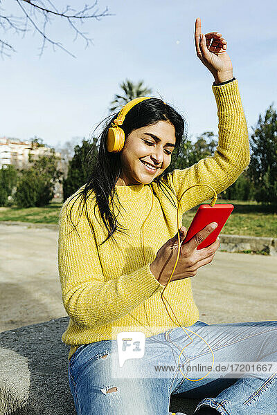 Lächelnde Frau mit Kopfhörern  die tanzt  während sie ein Handy im Park hält