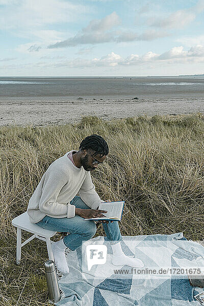 Junger Mann liest ein Buch und sitzt auf einem Hocker am Strand
