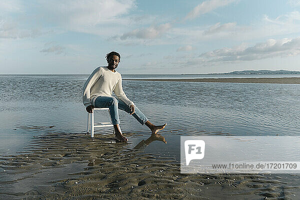 Lächelnder junger Mann sitzt auf einem weißen Hocker am Strand bei Sonnenuntergang