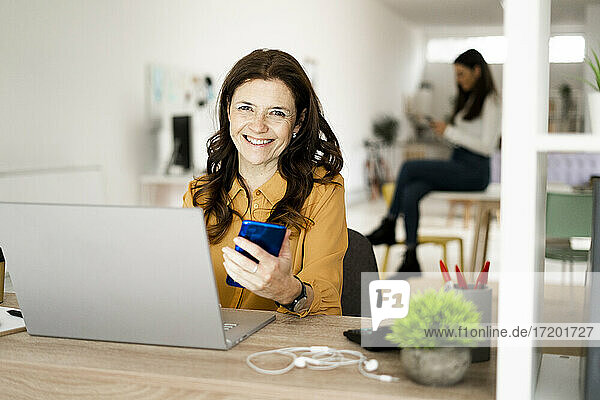 Lächelnde Geschäftsfrau mit Laptop  die ihr Smartphone hält  während sie an einem Schreibtisch im Heimbüro sitzt