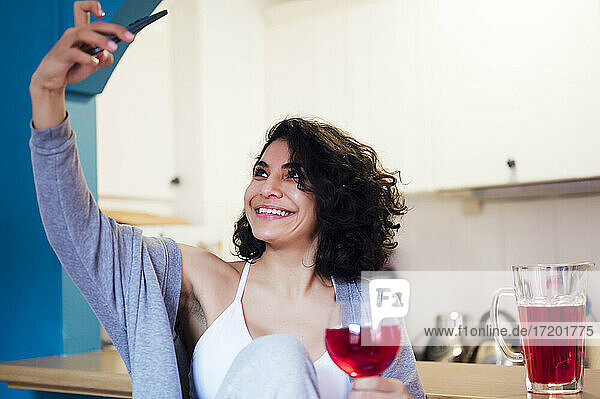 Lächelnde Frau hält ein Glas Saft und macht ein Selfie mit ihrem Handy in der Küche