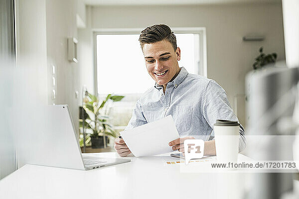 Lächelnder männlicher Fachmann  der ein Dokument liest  während er an einem Tisch im Heimbüro sitzt