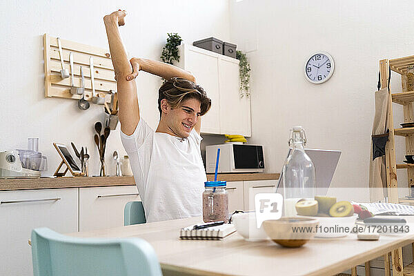 Young man stretching his hand while sitting at table with laptop in kitchen