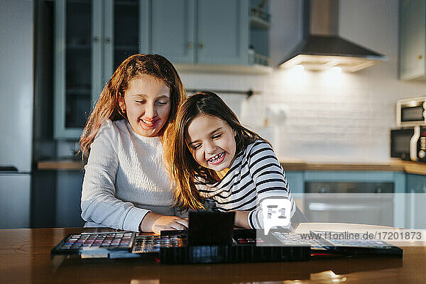 Fröhliche Mädchen spielen mit einer Lidschattenpalette  während sie am Esstisch in der Küche sitzen