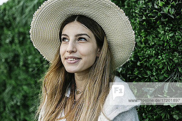 Nachdenkliche junge Frau mit Hut an einer grünen Pflanzenwand
