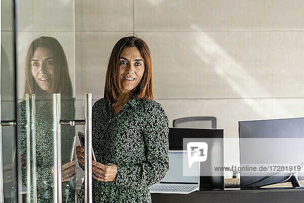 Lächelnde reife Geschäftsfrau mit Smartphone  die an einer Glastür in einer Kabine steht