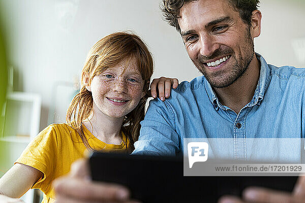 Lächelnder Vater und rothaariges Mädchen schauen auf ein digitales Tablet im Wohnzimmer