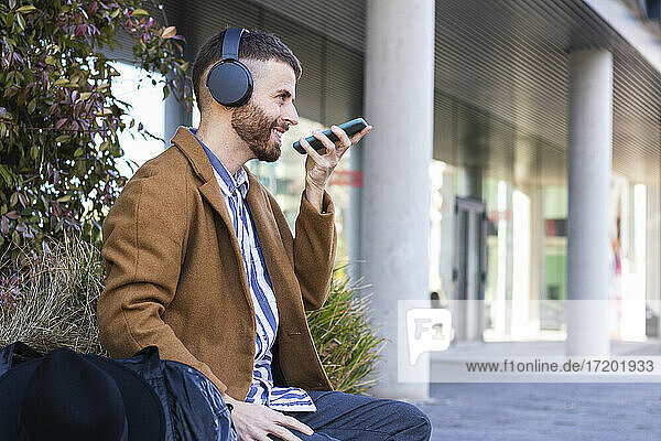Geschäftsmann mit Kopfhörern  der mit seinem Mobiltelefon spricht  während er mit einer Tasche im Freien sitzt