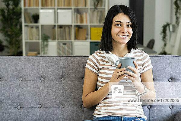 Glückliche Frau mit Kaffeetasse und kabellosen Kopfhörern auf dem Sofa zu Hause