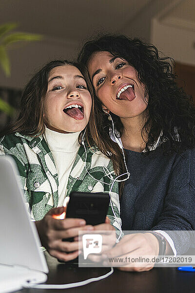 Mutter und Tochter mit Handy  das die Zunge herausstreckt  während sie zu Hause über Kopfhörer Musik hören