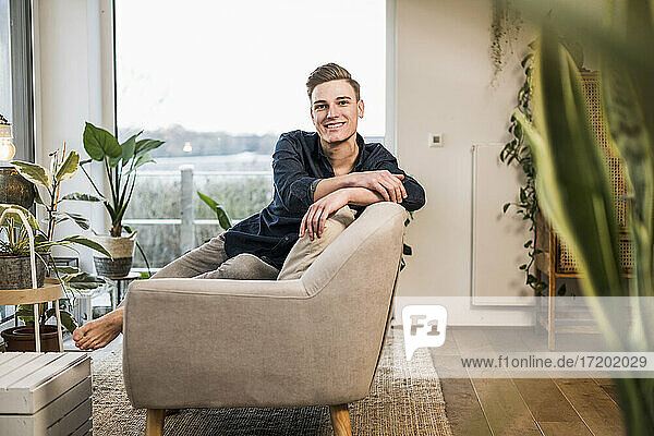 Lächelnder junger Mann sitzt auf dem Sofa vor dem Fenster im Wohnzimmer