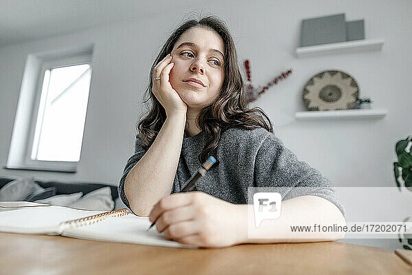Thoughtful woman sitting at table while writing in notebook in living room