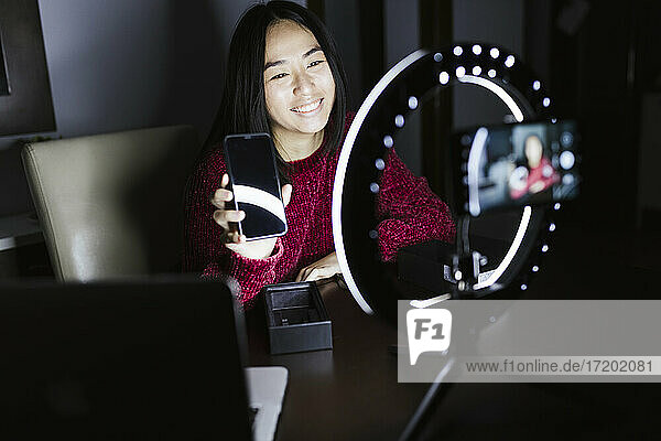 Lächelnde junge Influencerin hält ihr neues Smartphone in der Hand  während sie in der Dunkelkammer filmt