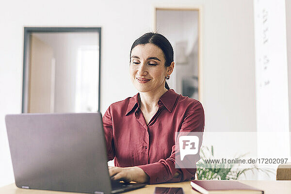 Lächelnde Unternehmerin  die einen Laptop benutzt  während sie am Tisch im Heimbüro sitzt