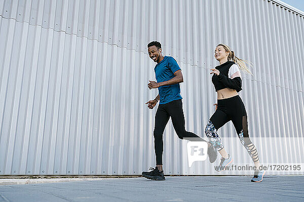 Smiling male and female athlete running on sidewalk by wall during sports training