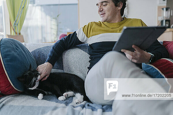 Smiling man with digital tablet stroking cat while sitting on sofa in living room